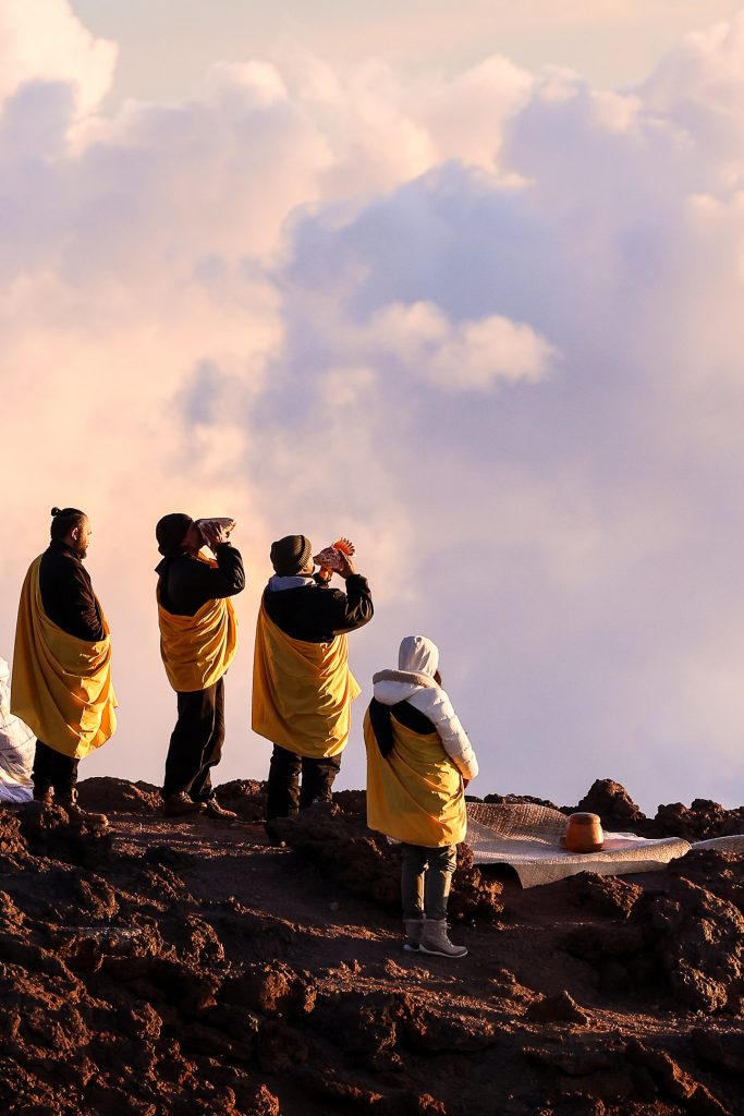 Four people on top of a mountain wearing black, white and yellow chanting at the sky